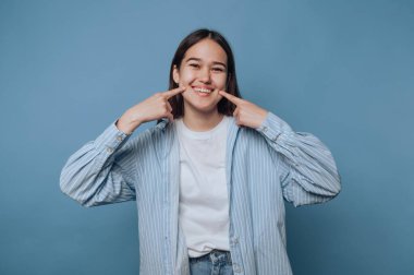 Genç bir kadın, parmaklarıyla dudaklarını işaret ederken neşeli bir şekilde gülümsüyor. O, sıradan bir kıyafet giymiş, yumuşak mavi bir arka planın önünde duruyor..
