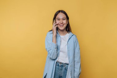 Genç bir kadın canlı sarı bir arka plana karşı duruyor, akıllı telefonuyla konuşurken geniş bir gülümsemeyle. Sıradan bir kıyafet giyiyor ve neşe saçıyor..