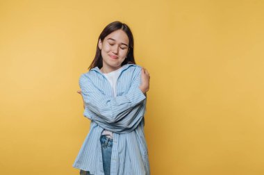 Genç bir kadın canlı sarı bir arkaplanın önünde duruyor. Açık mavi bir gömlek ve kot pantolon giyiyor, gülümsüyor ve kendini kucaklıyor, mutluluk ve kendini sevdiriyor..