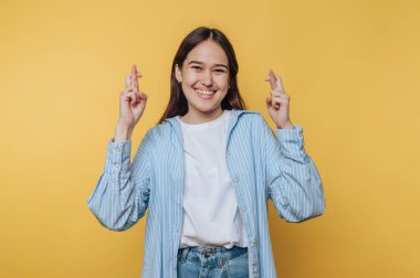 Genç bir kadın, parlak sarı bir duvarın önünde parmaklarını çaprazlarken mutlu bir şekilde gülümsüyor. Üzerinde mavi çizgili bir gömlek ve rahat bir kot pantolon var. Umudunu ve iyi şansını ifade ediyor..