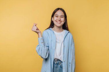 Neşeli genç bir kadın açık mavi çizgili bir gömlek ve kot içinde neşeli bir şekilde poz veriyor, eli ile canlı sarı bir arka plana karşı bir kalp hareketi yapıyor..