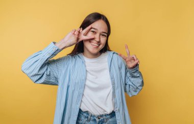 Genç bir kadın canlı sarı bir zemin üzerinde duruyor, elleriyle barış işaretleri yapıyor ve parlak bir şekilde gülümsüyor. Mavi çizgili gömlekli ve rahat kotlu sıradan bir kıyafet giyiyor..