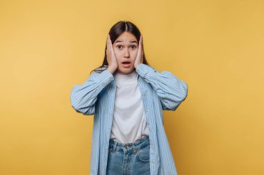 Genç bir kadın canlı sarı bir duvarın önünde duruyor, şaşkın şaşkın bakıyor ve elleriyle yüzünü tutuyor. Rahat bir kıyafet giyiyor, sıradan bir ortamda gerçek duygularını gösteriyor..