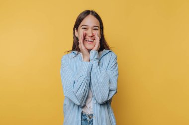 Elleri heyecandan yanaklarını sıvazlayan genç bir kadın geniş bir şekilde gülümsüyor. Canlı sarı bir arka planın önünde duruyor, rahat bir kıyafet giymiş, neşeli havasını sergiliyor..