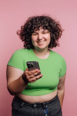 Kıvırcık saçlı neşeli bir genç, pembe bir arka fonun önünde duruyor. Akıllı bir telefon tutuyorlar ve gülümsüyorlar, rahat ve rahat bir tavır sergiliyorlar..