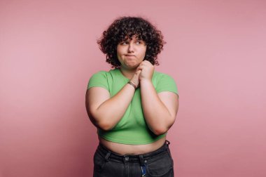 Genç bir birey, açık yeşil bluzu ve siyah pantolonu olan yumuşak pembe bir zemine karşı duruyor. Dalgın görünüyorlar, yüzlerinde ve ellerinde kıvırcık saçlar var..