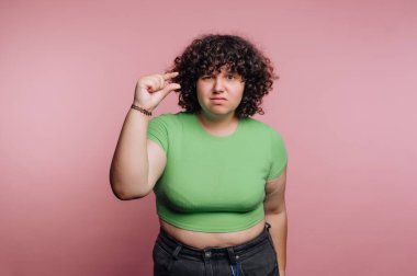 Pembe bir arka fonun önünde yeşil bluzu ve siyah pantolonu olan bir kadın duruyor. Küçük bir beden olduğunu göstermek için el hareketi yapıyor, neşeli bir tavır sergiliyor..