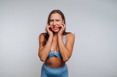 Bir kadın stüdyo ortamında mavi bir kıyafet giyerek yanaklarını tutarken endişeli bir yüz ifadesi sergiler. Sade arka plan duygularını geliştirir..