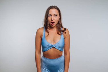 Mavi bir eşofman giymiş, düz bir arkaplanda duran bir kadın. İfadesi, şaşkınlığı geniş gözler ve hafif açık bir ağızla gösteriyor. Titrek bir an yaratıyor..