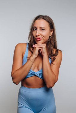 Parlak bir stüdyoda açık mavi bir bluz ve tayt giymiş genç bir kadın gülümserken çenesini şakaya vurarak dinlendiriyor. Kendine güveni ve tarzı dikkat çekiyor..