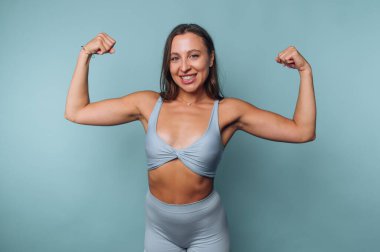 Bir kadın gücünü, açık mavi bir spor kıyafeti giyerken kollarını esneterek gösterir. Kameraya kendinden emin bir şekilde gülümsüyor, tonlu fiziğini vurguluyor..