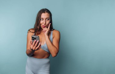 Bir kadın açık mavi bir duvara karşı duruyor, şık bir elbise giymiş. Elinde akıllı bir telefon tutuyor ve esprili bir şekilde poz veriyor. Bu da merak ya da sır olduğunu gösteriyor. İfadesi çekici ve hayat dolu..