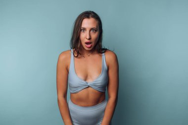 Açık mavi spor kıyafeti giyen bir kadın şaşkınlık ifadesi sergiliyor. Profesyonel bir stüdyo ortamında düz mavi bir arka plana karşı duruyor..