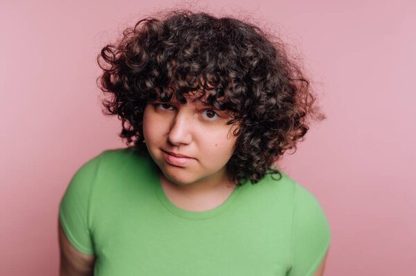 Молодой человек с вьющимися волосами стоит на мягком розовом фоне. Они носят ярко-зеленую рубашку и демонстрируют легкую ухмылку, источая чувство уверенности и стиля..