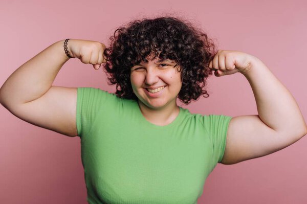 A young woman with curly hair smiles confidently while flexing her muscles. She wears a green shirt, and the pink backdrop highlights her cheerful expression, emphasizing empowerment.