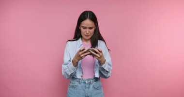Bir kadın pembe bir arka planın önünde duruyor, telefonuna bakarken şaşkınlık, şaşkınlık ve hayal kırıklığı gösteriyor. İfadesi bu anda hızla değişiyor..