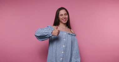 Bir kadın, parlak pembe bir ortamda, başparmakları ve neşeli ifadeleri de dahil olmak üzere çeşitli el hareketlerini sergiler. Parlak renkler neşesini arttırıyor, canlı bir atmosfer yaratıyor..