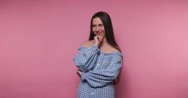 Genç bir kadın pembe bir arkaplana karşı duruyor, mavi kareli elbisesini gösteriyor. Düşünceli bir şekilde poz veriyor, gülümsüyor ve eğlenceli ve eğlenceli bir şekilde kameraya bağlanıyor..