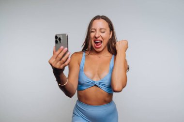 Genç bir kadın, mutluluğu ve heyecanı ifade eden bir selfie çekerken geniş bir gülümseme sergiliyor. Açık mavi bir elbise giymiş ve düz bir arkaplanda poz veriyor..