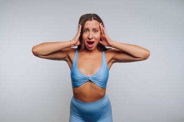 Genç bir kadın şaşırmış bir ifade sergiliyor, iki eliyle başını tutuyor. Mavi bir atletizm kıyafeti giyiyor ve düz bir zemine yaslanıp tepkisini vurguluyor..