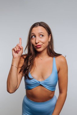 Genç bir kadın stüdyoda dikiliyor, mavi bir bluz giyiyor ve poposu uyumlu. Meraklı bir bakışla yukarıyı işaret ediyor, bir anlık bir farkındalık ya da fikir öneriyor..