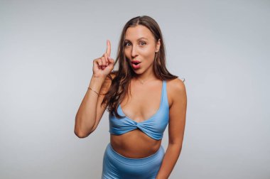 Genç bir kadın, açık renk bir stüdyoda mavi bir kıyafet giyerek dikiliyor. Bir parmağını kaldırdı ve şaşırmış bir ifadeyle enerji ve neşe dolu neşeli bir anı paylaştı..