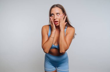 Mavi spor kıyafeti giyen bir kadın şaşırmış ve düşünceli görünüyor. Bir ışık arkaplanına karşı poz veriyor, ifadesine ve vücut diline neşeli bir dokunuş ekliyor..