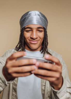 Genç bir adam akıllı telefonuna odaklanmış, oyun oynarken gülümsüyor. Başörtüsü takıyor ve evin içinde rahat bir ortamda oturuyor. Hafif ışıklandırmayla aydınlatılıyor..