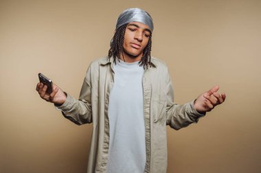 Örgülü saçı olan genç bir birey evde duruyor, bir elinde akıllı bir telefon tutarken şaşkın bir ifade sergiliyor. Ayarlama düz, nötr bir arkaplan içeriyor.