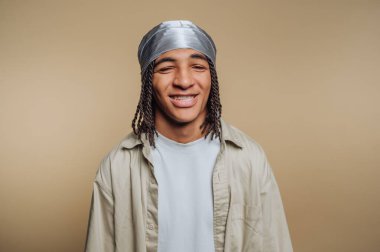 Genç bir adam büyük bir gülümsemeyle özgüvenle ayakta duruyor. Saçlarını ördü ve açık gömlekli gümüş bir eşarp takıyor. Arka plan onun ifadesini vurgulayan yumuşak, nötr bir ton..