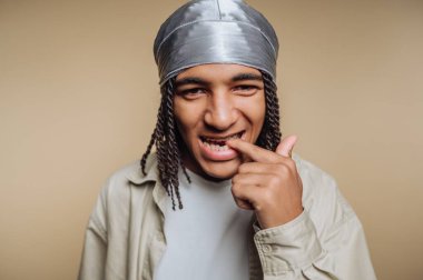 Örgülü saçları ve gümüş başörtüsü olan genç bir adam gülümsüyor ve şakacı bir şekilde dişlerine dokunuyor. Tarafsız bir arkaplanda poz veriyor, içeride eğlenceli bir an sergiliyor..
