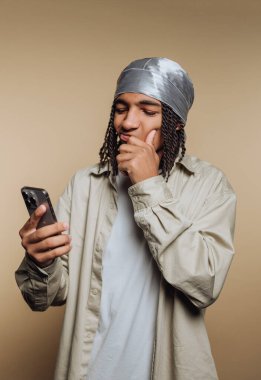 Gümüş bir başörtüsü ve açık renk bir gömlek giyen genç bir adam akıllı telefonunu kontrol ederken derin düşüncelere dalıyor. Tarafsız arka plan, ifadesine odaklanmasını sağlıyor..