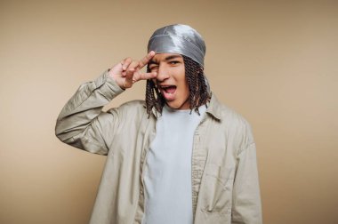 Gümüş bandana takan genç bir adam kameraya gülümserken eliyle eğlenceli bir jest yapar. Açık renk bir gömlek giyiyor ve saçları örgülü..