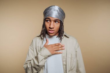 Elleri göğsünde, şaşırmış bir şekilde duran genç bir insan. Gri bir başlık ve açık renkli bir kıyafet giyiyorlar. Çevrelerinde ifadelerini vurgulayan tarafsız bir zemin var..