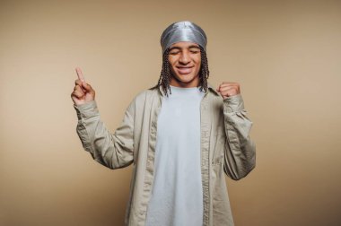 Genç bir insan büyük bir gülümsemeyle kollarını kaldırıyor. Basit bir stüdyoda gümüş bir saç bandı ve rahat bir kıyafet giyiyorlar..