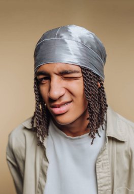 Genç bir adam başörtüsü takarken neşeyle göz kırpıyor. Zarif rastaları, yumuşak, tarafsız bir arka plana karşı kendine güvenen bir duruş sergilerken yüzünü çerçeveliyor..