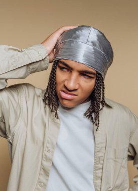 Örgülü saçlı genç bir adam gümüş bir başörtüsü ve açık renk bir gömlek giyiyor. Kafasına dokunurken kafası karışmış görünüyor, yumuşak bir bej arka plana yaslanmış..
