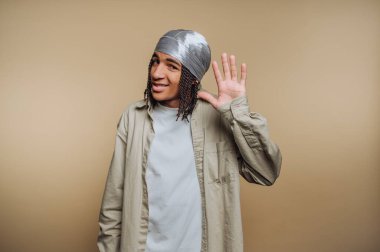 Genç bir adam dostça bir gülümsemeyle duruyor, neşeli bir hareketle elini kaldırıyor. Hafif bir gömlek ve türban giyiyor. Basit bir arkaplanı var..