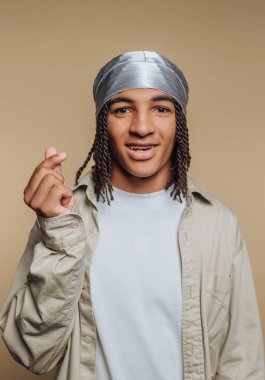 Genç bir adam kendine güvenerek duruyor, gümüş bir başörtüsü ve açık renk bir gömlek giyiyor. Elleriyle bir kalp hareketi yapıyor. Tarafsız bir ortamda pozitiflik ve cazibe yayıyor..