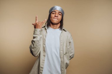 Genç bir adam kendine güvenerek poz veriyor, gümüş bir bandana ve açık renk elbiseler giyiyor. Stüdyoda nötr bir arkaplan karşısında dururken el işareti yapıyor..