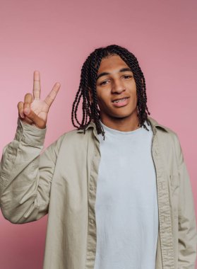 Genç bir adam, örgülerini ve günlük kıyafetlerini gösteren bir barış işaretiyle güvenle ayakta duruyor. Kameraya gülümsüyor, yumuşak pembe bir arka plana karşı dostane bir ortam yaratıyor..