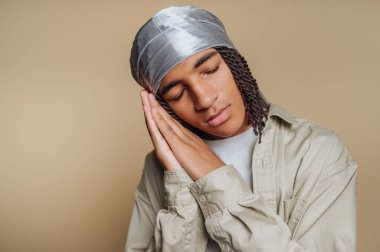 Genç bir adam gözleri kapalı dinleniyor, huzurlu bir ifade sergiliyor. Örgülü bir saç stili ve başörtüsü takıyor. Ev ortamında sakin bir hava yaratıyor..