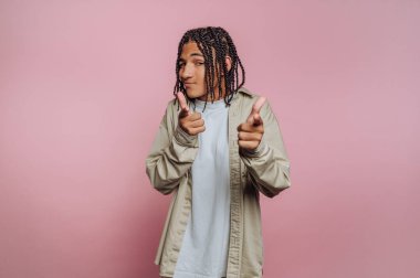 Genç bir insan pembe bir arkaplana karşı duruyor, kendine güvenerek poz veriyor ve parmaklarını şakaya vuruyor. Günlük kıyafetler giyiyorlar ve saçlarını ördürüp, eğlenceli bir hava yayıyorlar..