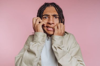 Örgülü saçları olan genç bir adam yanaklarını tutarak şok olmuş bir ifade sergiliyor. Arka plan yumuşak bir pembe, etrafında neşeli ve canlı bir atmosfer yaratıyor..