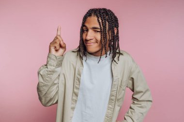 Genç bir adam örgülü saç stilini neşeli bir ifadeyle sergiliyor. Pembe bir arka fonun önünde güvenle duruyor, sanki bir fikri paylaşıyormuş gibi yukarı bakıyor..