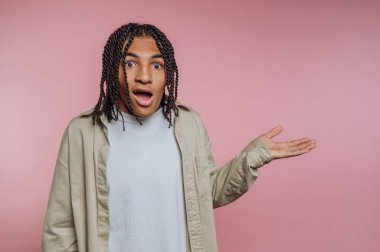 Örgülü saçlı genç bir adam pembe bir arka plana karşı duruyor, tek eliyle hareket ederken şok olmuş bir ifade sergiliyor. Sahne, sakin bir atmosferde bir sürpriz anı yakalıyor..