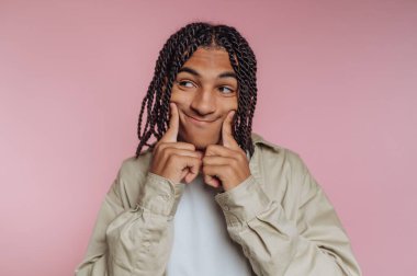 Genç bir adam, komik bir yüz ifadesi takınarak, yanaklarına dokunarak geniş bir gülümseme sergiliyor. Canlı pembe arka plan, sahneye canlı bir dokunuş katıyor..