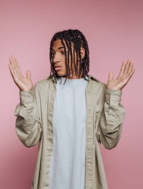 Elleri havada duran genç bir insan, yumuşak pembe bir arka plana karşı gevşek bir yüz ifadesi sergiliyor. Sahne, kişisel ifade ve tarzın bir anını yakalar.