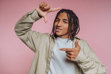 Açık renk gömlek giymiş genç bir adam yumuşak pembe bir arka plana karşı neşeli bir poz veriyor. Örgülü saç stili kendine güvenini arttırıyor. Parmak hareketleriyle yaratıcılığını gösteriyor..