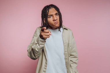 Genç bir adam pembe bir arkaplana karşı durarak kameraya ciddi bir ifadeyle bakıyor. Örgülü saçı çarpıcı görünüşüne ekleniyor..